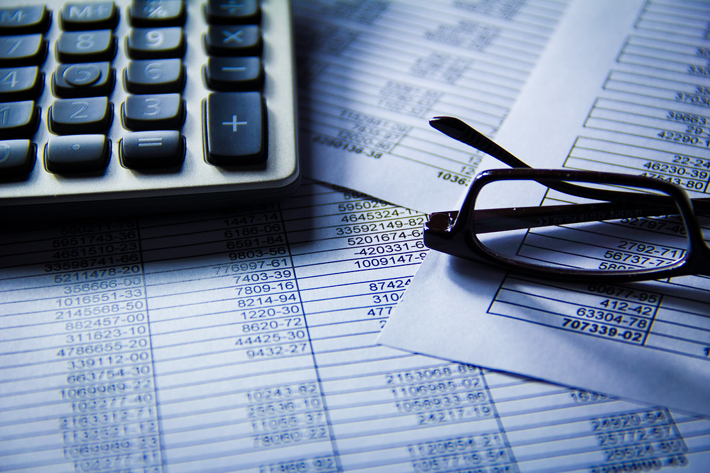 Gros plan sur une calculatrice de bureau et une paire de lunettes noires posées sur des documents remplis de tableaux de chiffres financiers.